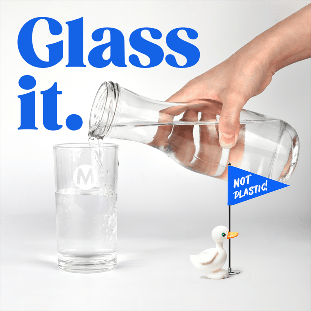 Bicchiere di vetro riempito di acqua da una bottiglia di vetro e un pupazzo di papero che sventola la bandiera NOT PLASTIC
