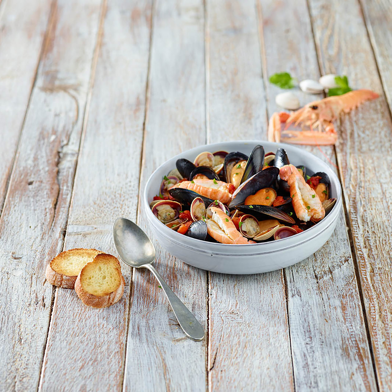 Piatto di cozze, vongole e scampi posizionato vicino a un cucchiaio, due crostini di pane e davanti a uno scampo e a delle vongole. 